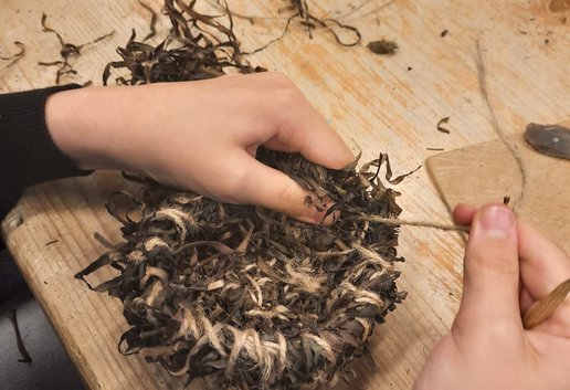 Auf dem Foto ist zu sehen wie ein Körbchen aus Seegras genäht wird. 