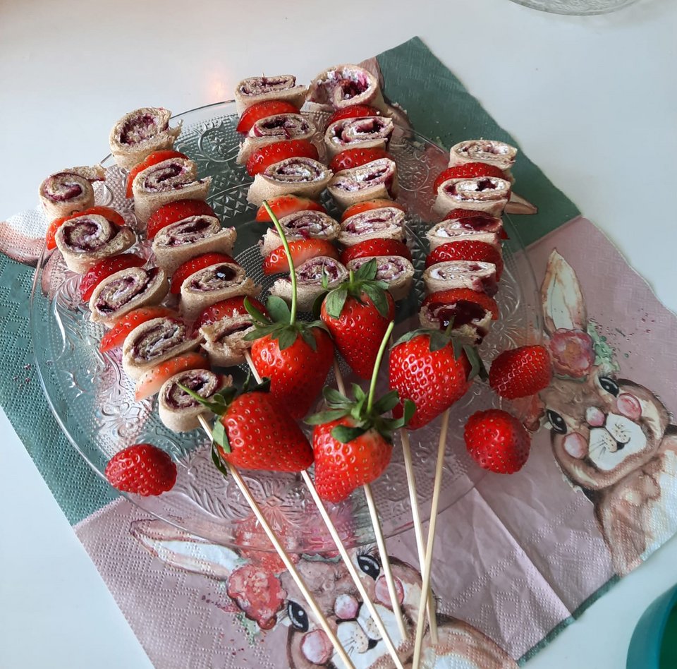Fünf Holzspieße mit aufgerollten Pfannkuchenstreifen und Erdbeeren im Wechsel aufgesteckt liegen auf einem Glasteller. Der Glasteller steht auf einer bunten Serviette mit Hasenmotiven. Einzelne Erdbeeren liegen als Dekoration auf dem Teller.