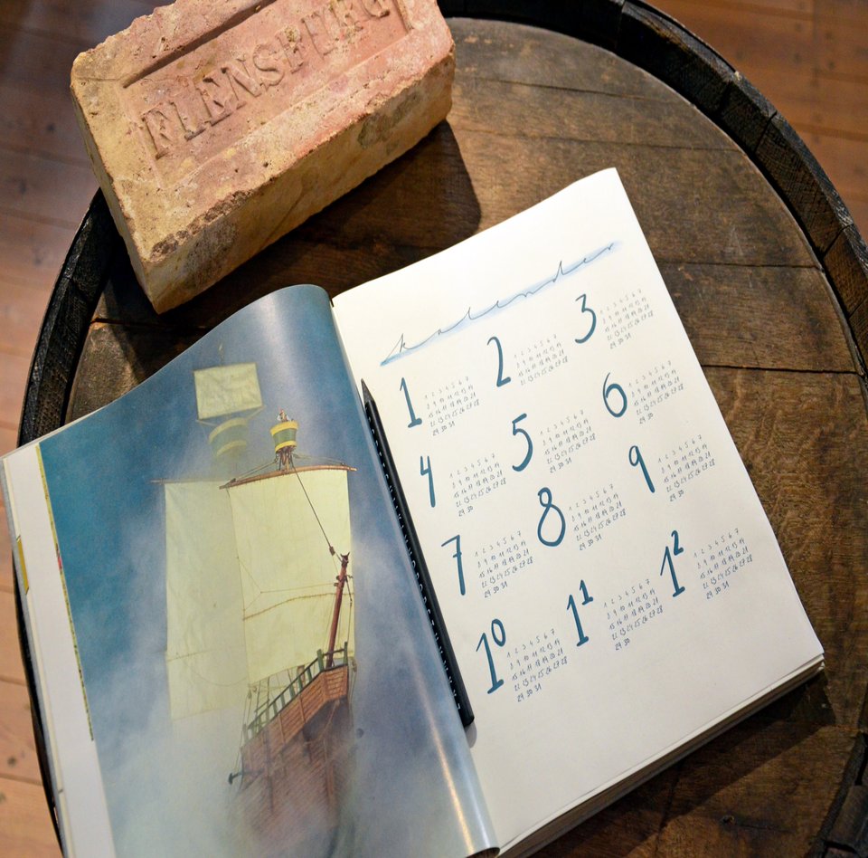 Auf einem Fass liegt ein Terminkalender. Dieser ist aufgeschlagen und eine Jahresübersicht ist zu sehen. Daneben liegt ein Ziegelstein mit dem Aufdruck „Flensburg“.