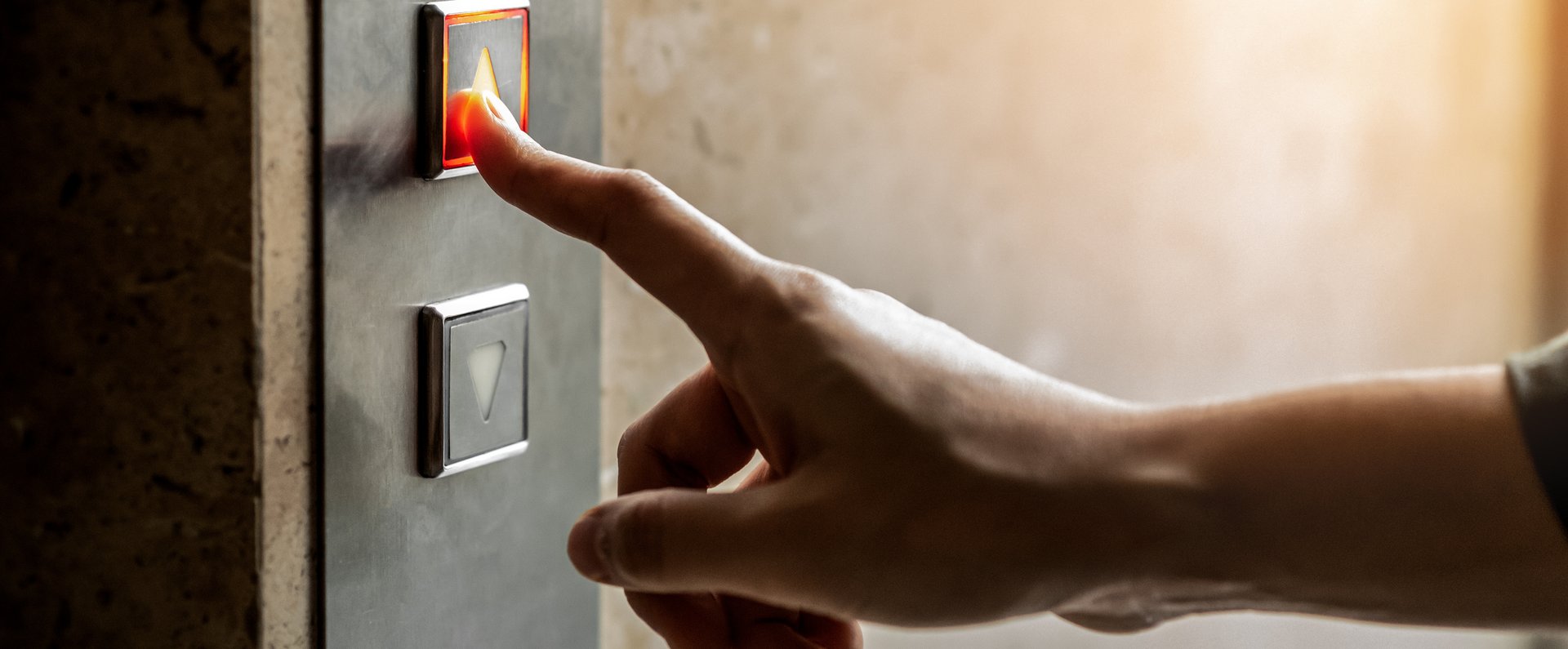 Nærbillede af en hånd. På en væg ses en elevators betjeningspanel. Den har to knapper med hver en pil. Den ene peger opad, den anden nedad. Hånden er strakt ud mod panelet. Pegefingeren rører den øverste knap.
