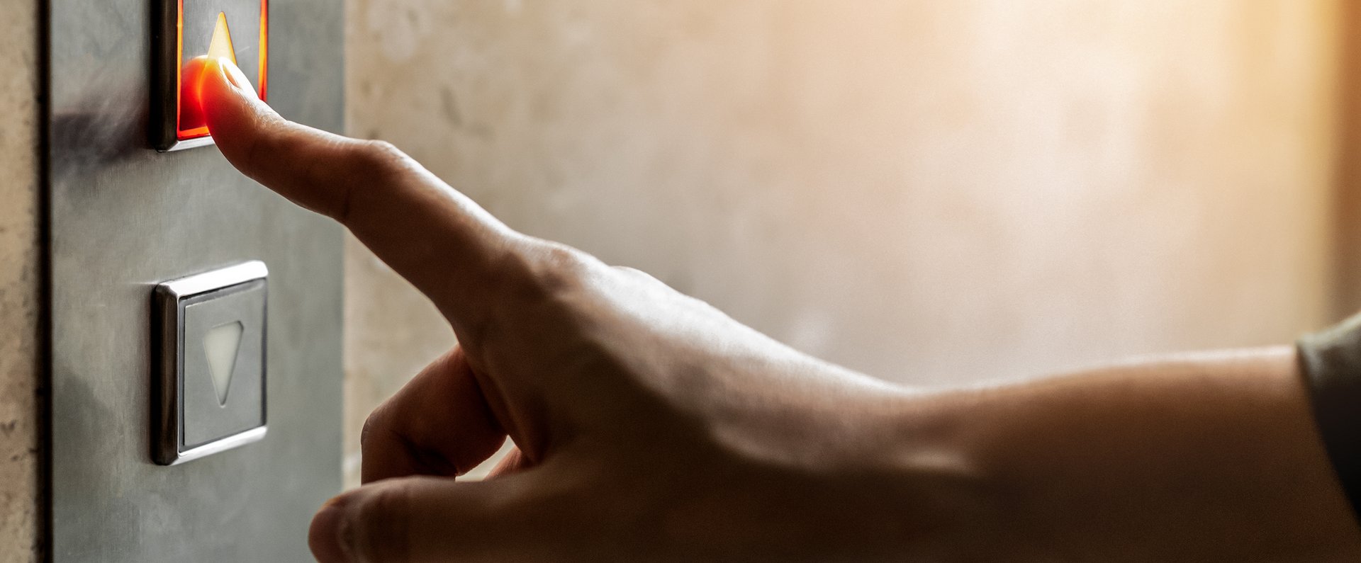 Nahaufnahme von einer Hand. An einer Wand ist das Schaltpanel eines Aufzuges zu sehen. Dieses hat zwei Knöpfe mit jeweils einem Pfeil. Der eine zeigt nach oben, der andere nach unten. Die Hand ist in Richtung dieses Panels ausgestreckt. Der Zeigefinger berührt den oberen Knopf.