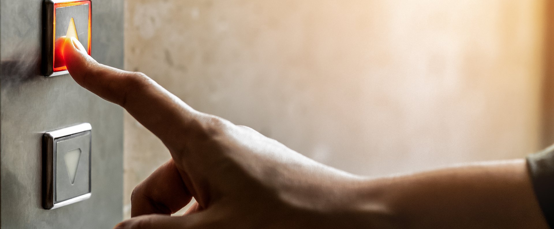 Nahaufnahme von einer Hand. An einer Wand ist das Schaltpanel eines Aufzuges zu sehen. Dieses hat zwei Knöpfe mit jeweils einem Pfeil. Der eine zeigt nach oben, der andere nach unten. Die Hand ist in Richtung dieses Panels ausgestreckt. Der Zeigefinger berührt den oberen Knopf.