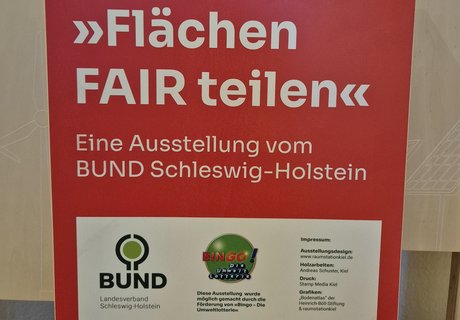 Das Foto zeigt einen Holzaufsteller mit Texttafel. Auf der Texttafel sind der Name der Wanderausstellung sowie die Gestalter als auch Sponsoren abgebildet. 